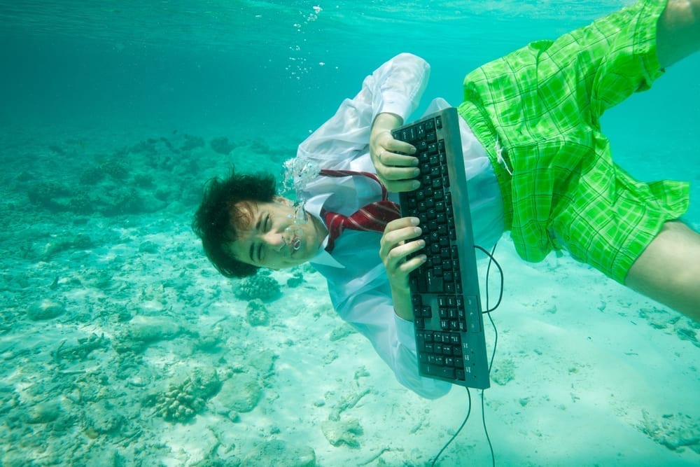 A person in a shirt and tie and shorts under the water in the sea with a computer keyboard