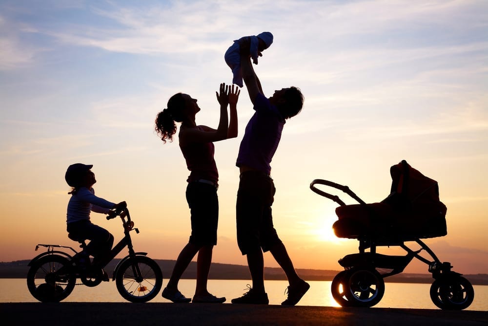 Photo of parents holding a baby aloft with a child on a bike and a pushchair at the side